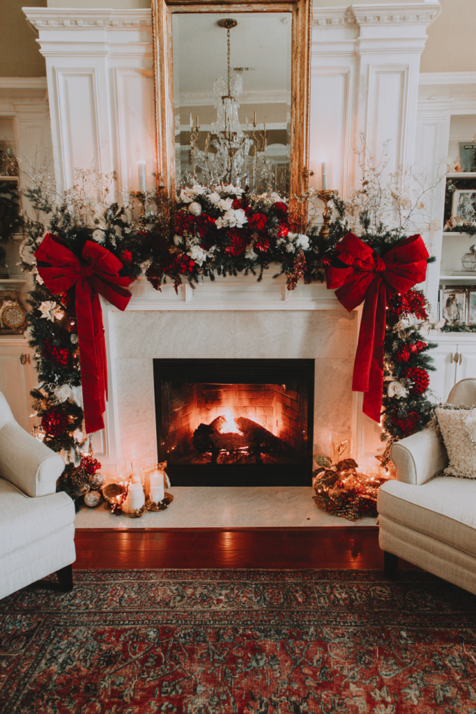 A grand white fireplace decorated for Christmas featuring a large gold mirror and a thick garland filled with greenery, white flowers, and red blooms. Two massive, oversized red velvet bows hang symmetrically on either side of the mantel, trailing down towards the floor. A warm fire burns in the hearth, adding a cozy glow to the room.